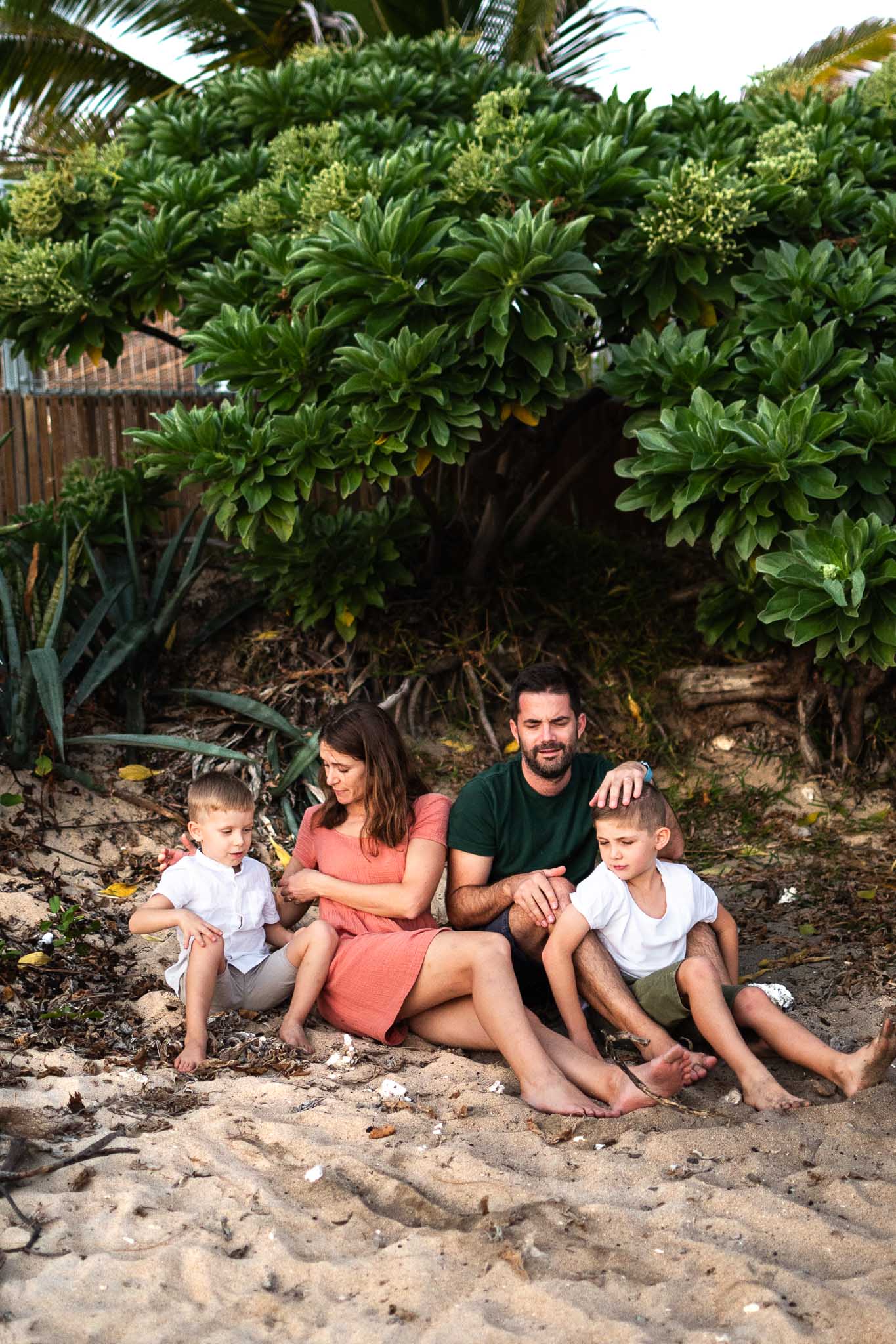 une famille est assise sur le sable devant un arbustre